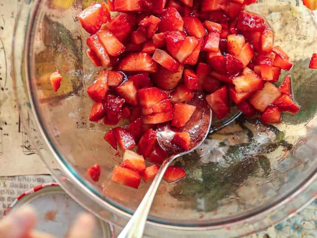 A glass bowl filled with diced strawberries. A silver spoon is partially submerged in the fruit mix. The background includes a tabletop with a decorative paper and a jar lid next to the bowl.